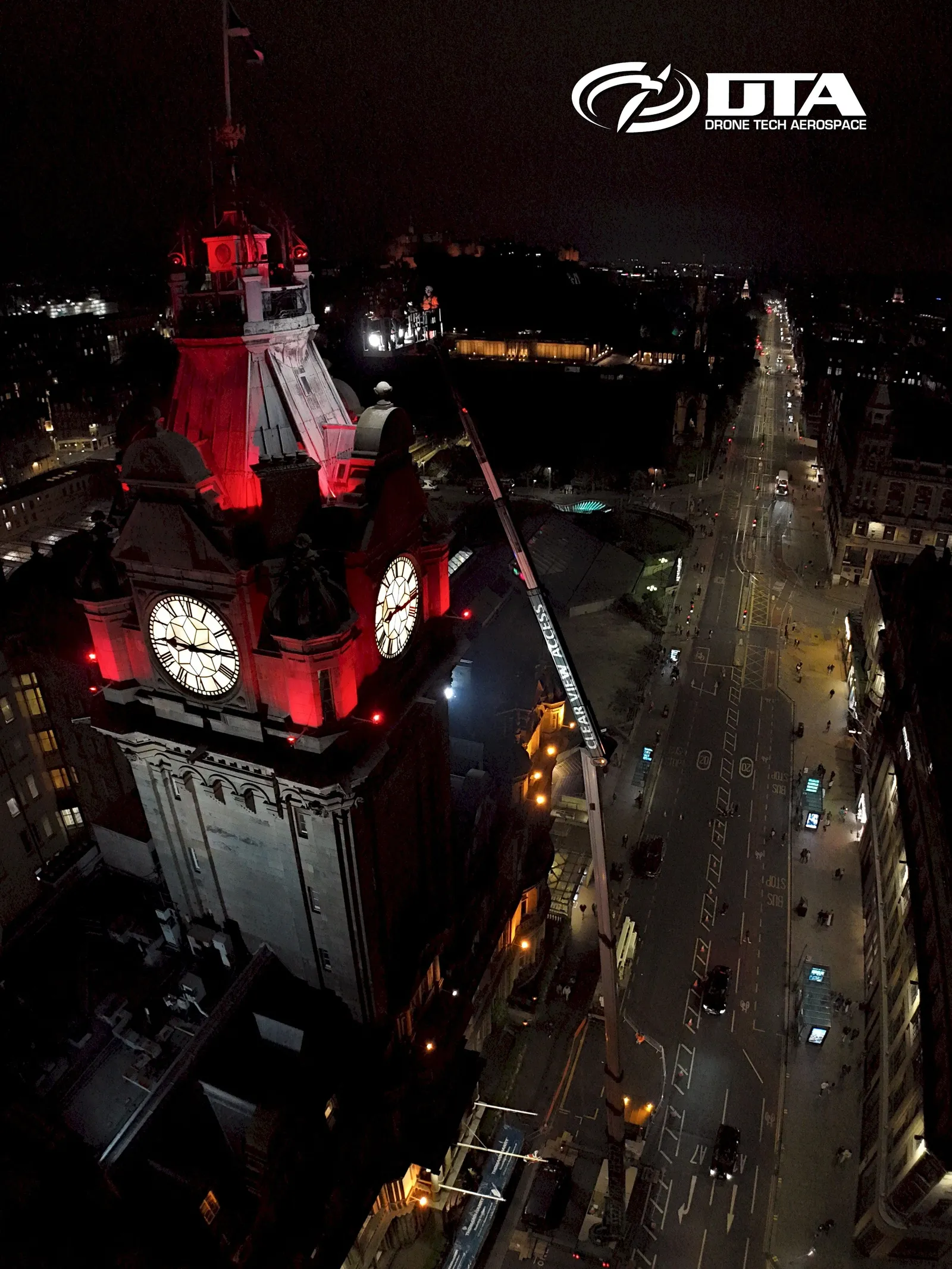 Vertical nighttime aerial photograph of an illuminated clocktower above a city street lined with lights.