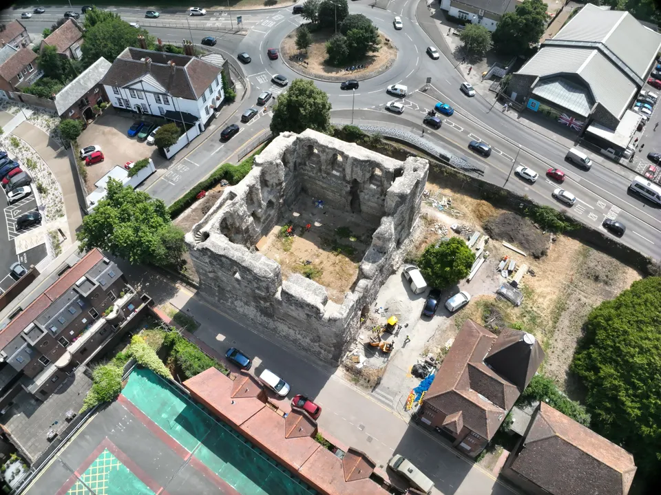 Straight-down aerial photograph of a roofless square stone keep with thick ruined walls, workers and materials inside, beside a busy road junction.