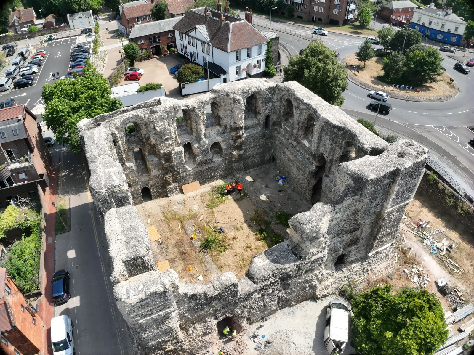 Oblique aerial photograph of a ruined square stone keep with arched openings in its inner walls, surrounded by streets, houses and trees.