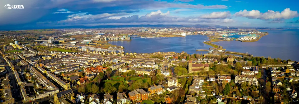 Wide panoramic aerial photograph of a waterfront bay with a barrage and surrounding buildings.