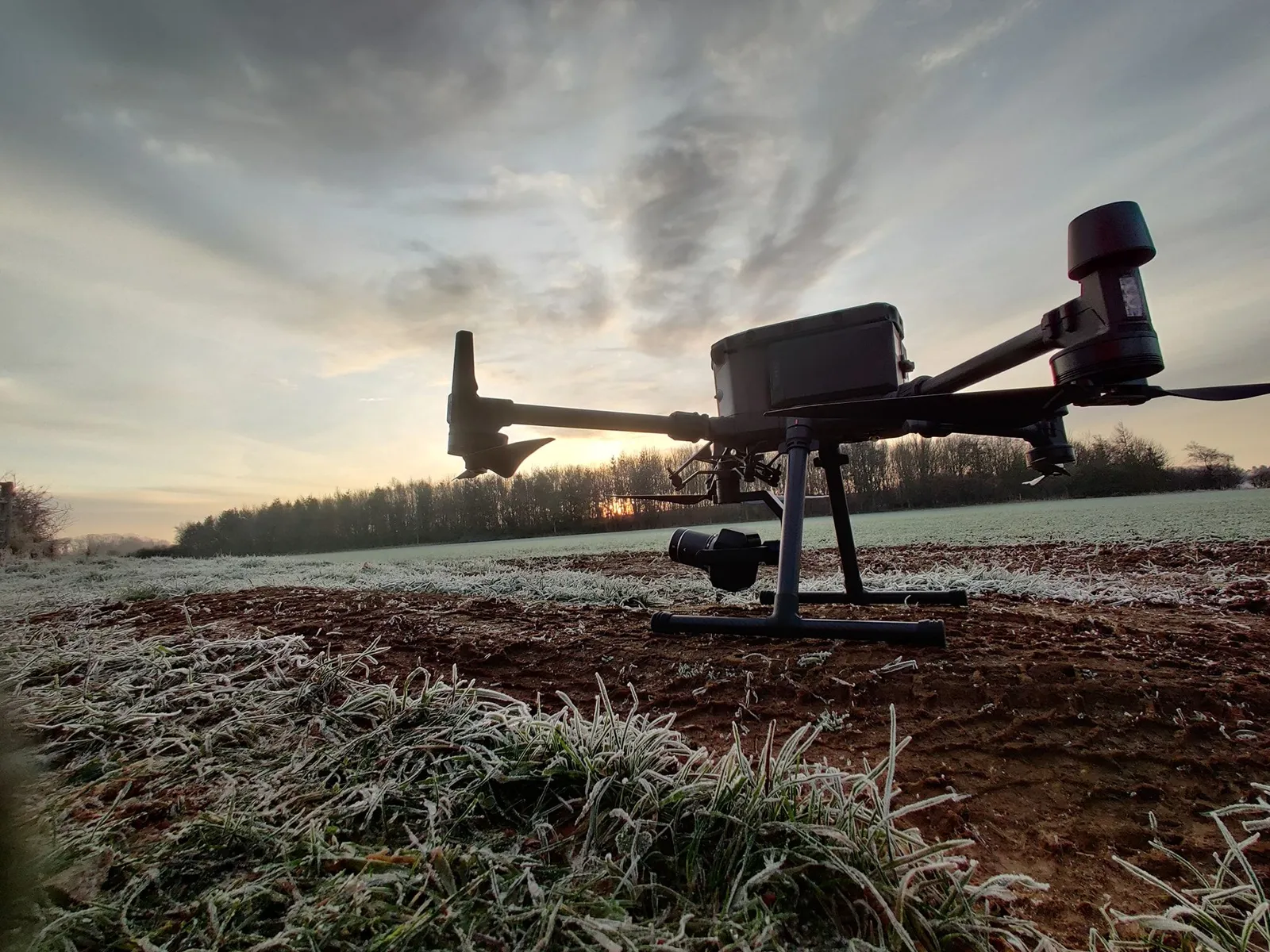 Low-angle photograph of a large grey survey drone standing on frosted ploughed ground with a sunrise and a line of trees behind it.