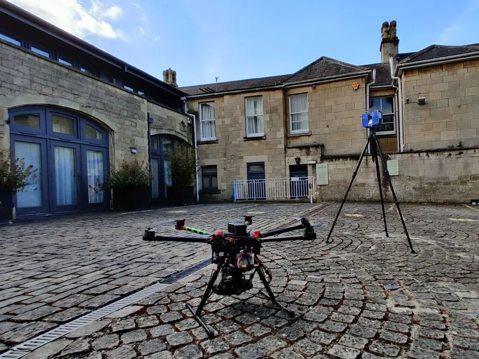 Photograph of a drone resting on the ground beside a tripod-mounted laser scanner.