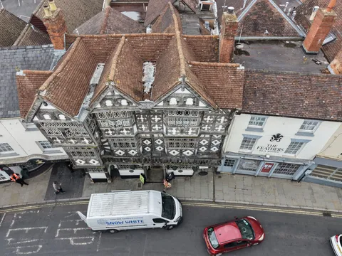 Aerial view straight down onto clay tiled roofs, brick chimneys and black and white timber framed frontages on a Ludlow street, with a van and cars below.