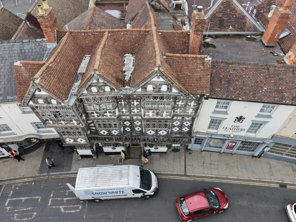 Aerial view straight down onto clay tiled roofs, brick chimneys and black and white timber framed frontages on a Ludlow street, with a van and cars below.