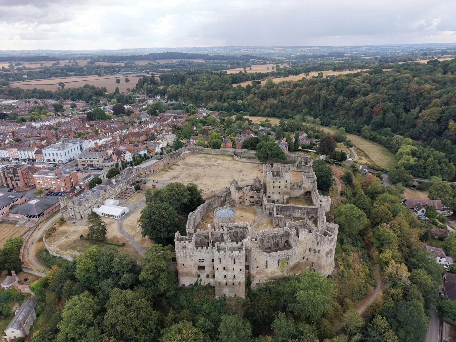 Aerial view of a ruined stone castle with a large dry outer bailey, surrounded by trees, with a market town and farmland behind.