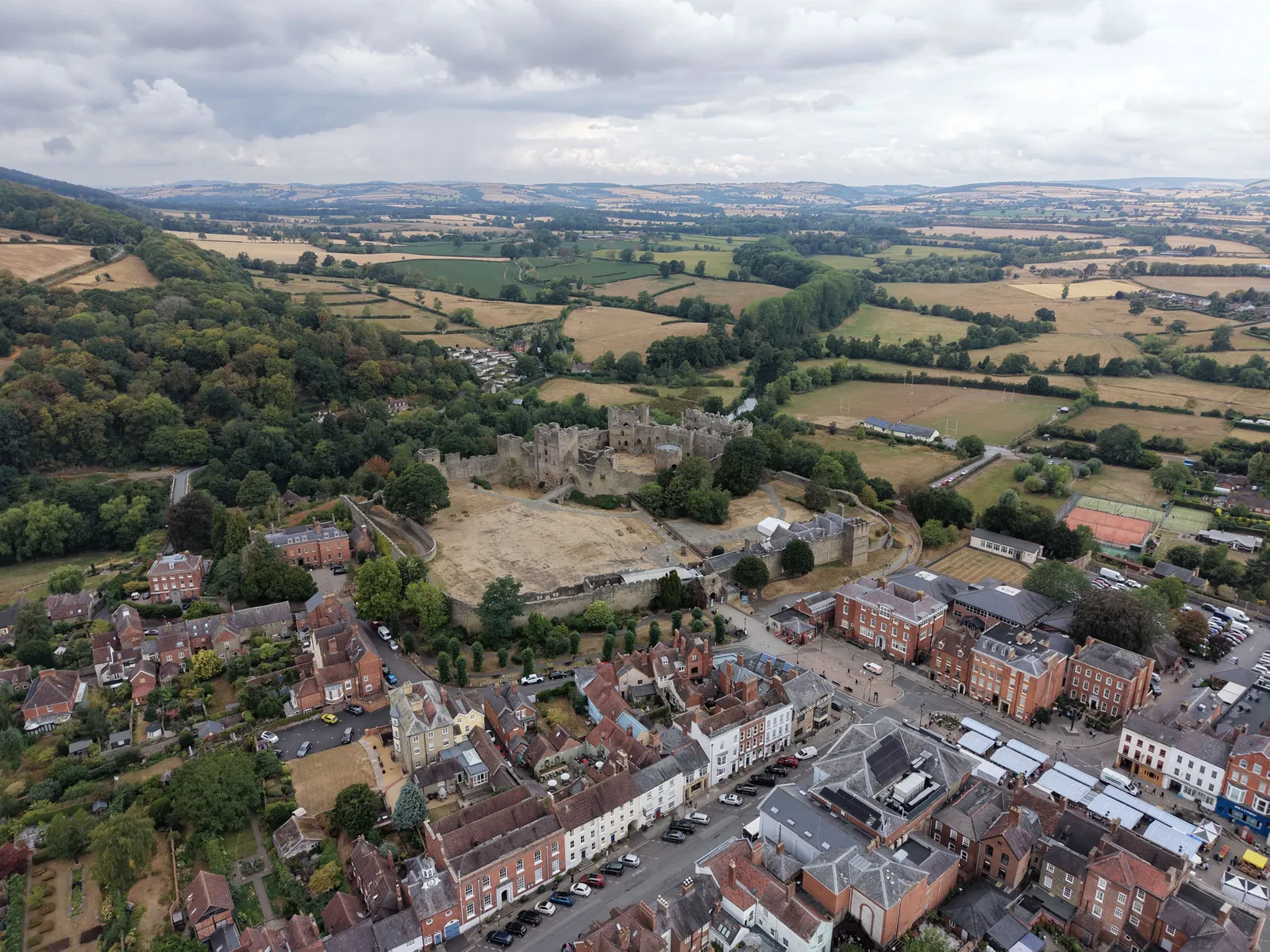 High aerial view of a ruined castle beside a market square and rows of red brick and rendered town houses, with fields and wooded hills beyond.