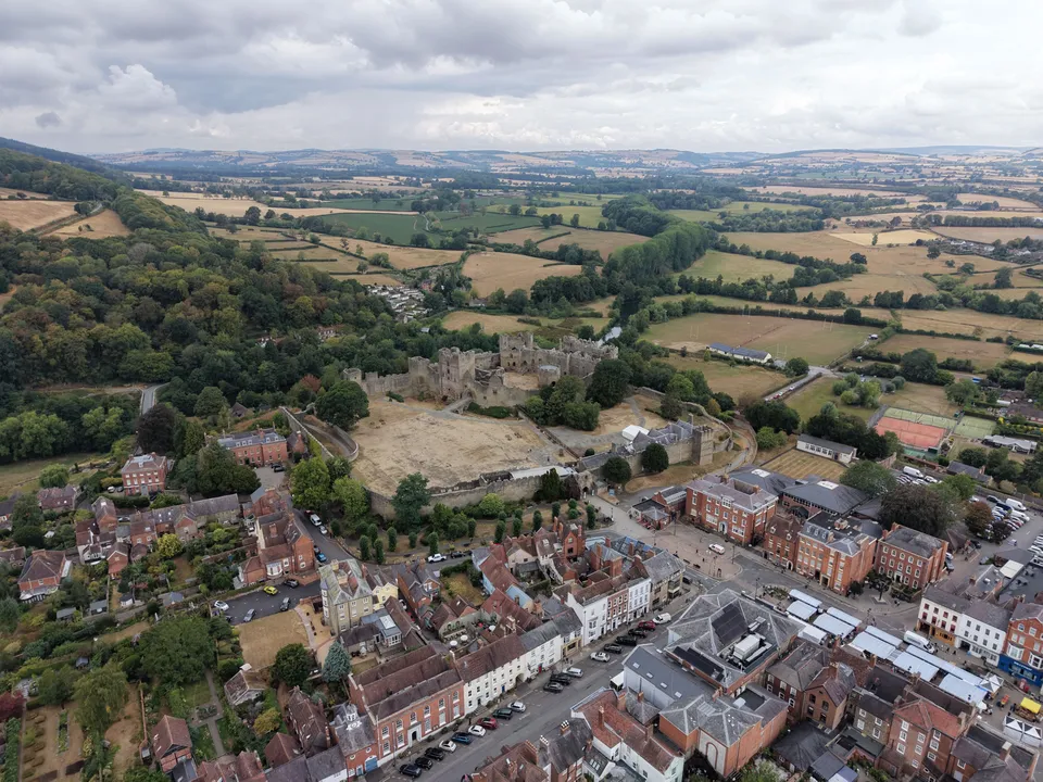 High aerial view of a ruined castle beside a market square and rows of red brick and rendered town houses, with fields and wooded hills beyond.
