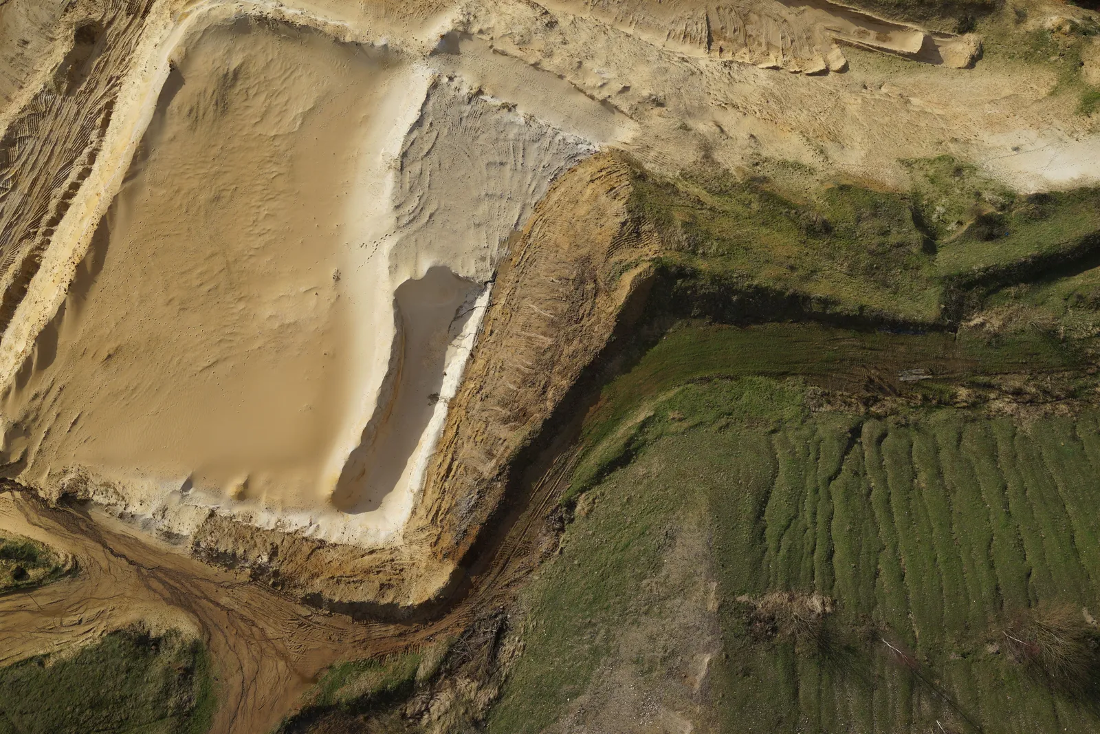 Straight-down aerial photograph of a pale sand extraction cell bounded by a brown overburden bank, with a haul road and rough grassland alongside.