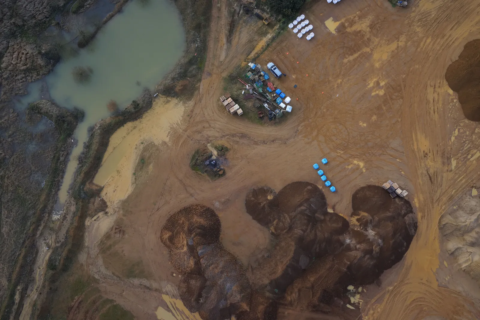 Straight-down aerial photograph of dark stockpiles, a sandy yard with parked plant and stacked bags, and a green silt lagoon at the top left.