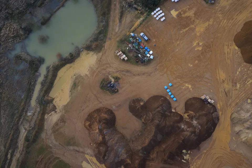 Straight-down aerial photograph of dark stockpiles, a sandy yard with parked plant and stacked bags, and a green silt lagoon at the top left.