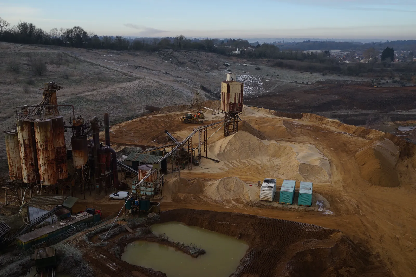 Aerial photograph of a sand and gravel plant with rusted silos, a conveyor, pale stockpiles, a muddy pond and frost on the surrounding ground at sunrise.