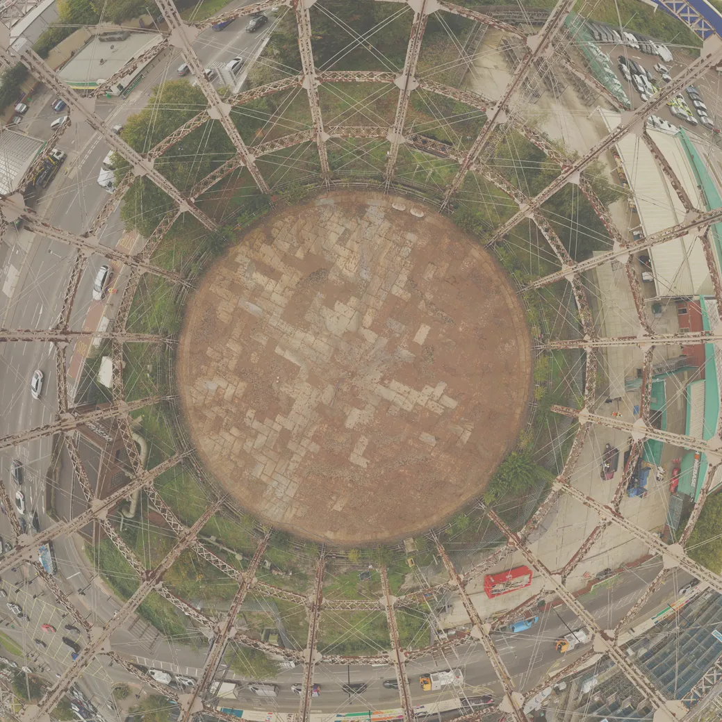 Fisheye aerial photograph looking straight down into a derelict circular gas holder structure surrounded by dense urban buildings.