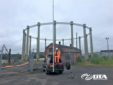 4x4-mounted elevated MAST raised beside a vehicle at an industrial site with historic gasholder framework behind