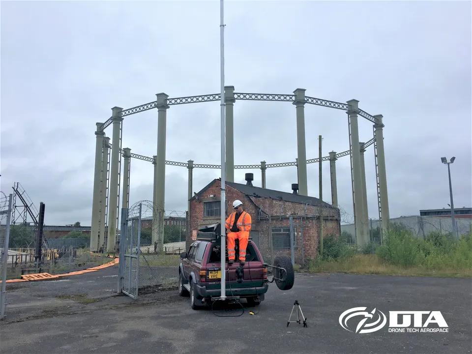 4x4-mounted elevated MAST raised beside a vehicle at an industrial site with historic gasholder framework behind
