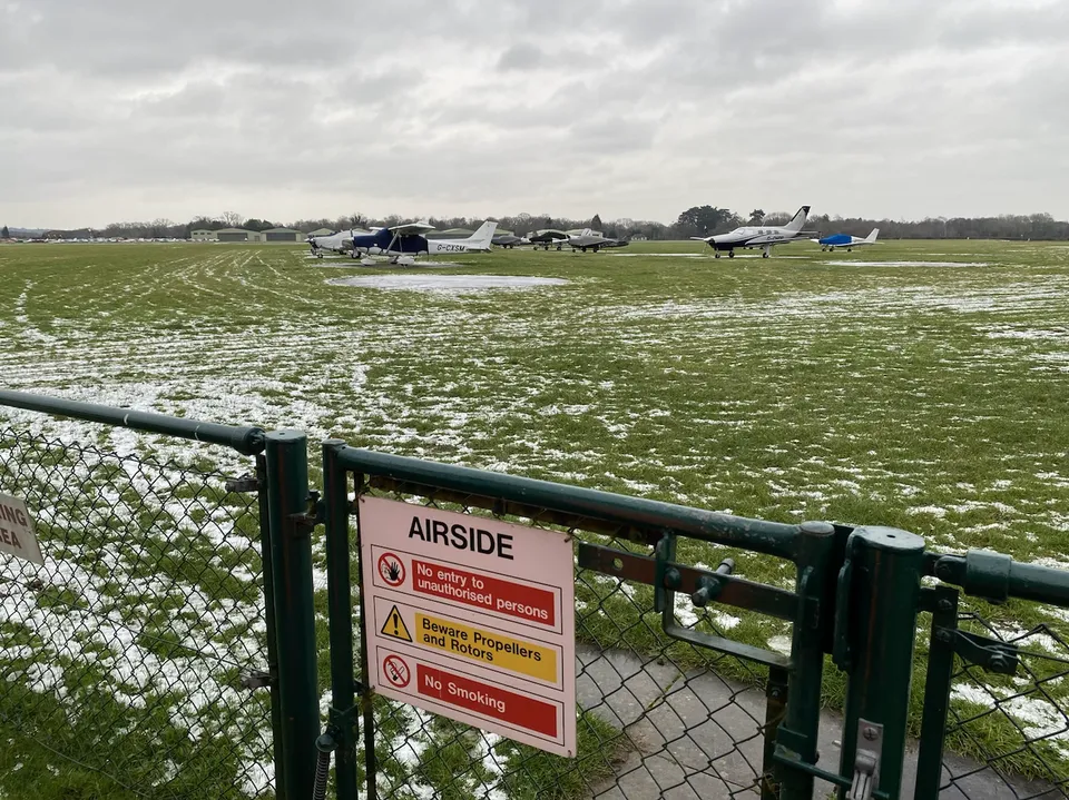 Airside access gate at Denham Aerodrome with light aircraft parked on the grass apron beyond