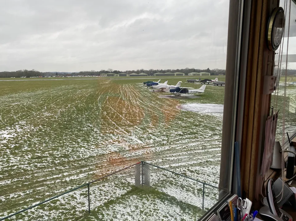 View from inside Denham's air traffic control tower across the apron toward parked light aircraft and hangars