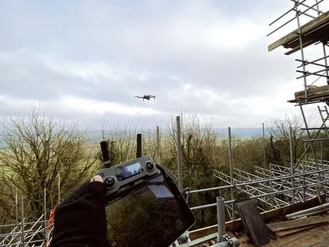 Drone in flight beside full scaffolding on a historic building during a structural survey, with rolling countryside beyond