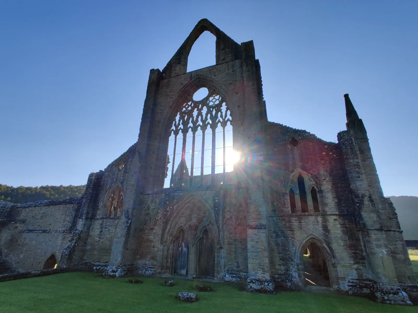 Tintern Abbey's ruined Gothic west window and facade with sun flaring through the stone tracery against a clear blue sky